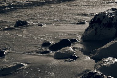 Düz kayalar kumlu kıyı şeridini noktalarken, sakin bir gündoğumu sırasında yumuşak dalgalar sahile çarpıyor ve huzurlu bir sahil atmosferi yaratıyor..