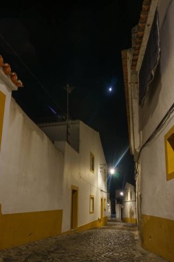Gökyüzünde ay, Evora, Alentejo, Portekiz 'de gece vakti sıcacık beyaz ve sarı evleri olan Evora' nın dar asfalt sokak manzarası.