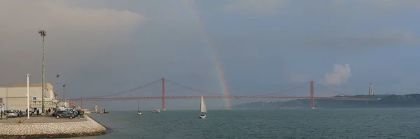 Panorama of rainbow over red bridge 25 de Abril Bridge with sailing boat during sunset, Lisbon, Portugal