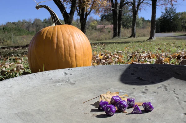 Close up of purple role playing game dice on stone ruins with a pumpkin and autumn countryside in the background.