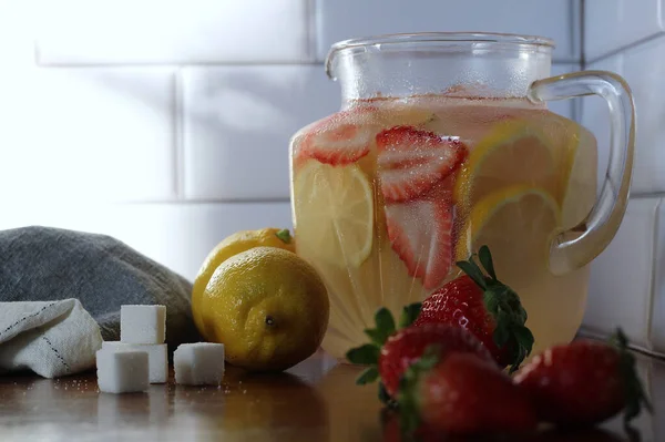 Close up of strawberry lemonade with sugar cubes, lemons, strawberries, and a kitchen towel.