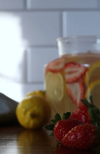 Close up of two strawberries with strawberry lemonade, lemons, and a kitchen towel blurred in the background.