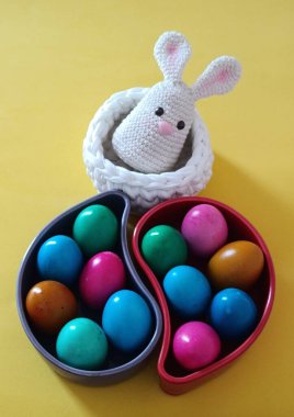 Bayramlık Paskalya temalı kompozisyon. Küçük, beyaz bir örgü sepetinin içine yerleştirilmiş küçük beyaz bir örgü tavşanı. Her biri renkli paskalya yumurtalarıyla dolu iki gözyaşı damlası kabı var.