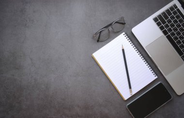 Düz görünüm dizüstü bilgisayarı ve cep telefonu açık siyah ofis masasında defter, kalem ve gözlüklerle yatıyordu.