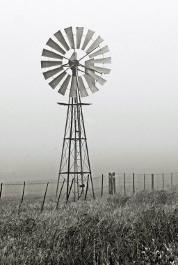 Yel değirmeni su pompasıyla manzara ve sabah sisli tek renkli bir kasaba yolu.