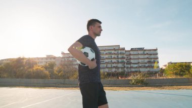 Kolunun altında bir futbol topuyla yürüyor..