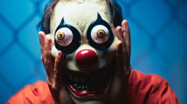 Korkunç maske takan, sahte göz küreleri olan, ellerini yüzüne bastıran, turuncu takım elbiseli, mavi arka planlı bir palyaço..