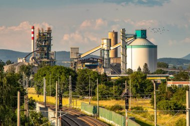 Ön planda fabrika bacası ve demiryolu hattı olan bir çimento fabrikasının dışı..