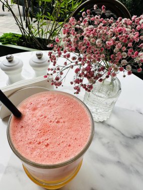 Bir bardak taze sıkılmış meyve suyu ve açık bir kafedeki masada duran pembe minyatür çiçeklerden oluşan bir vazo yakın plan.. 