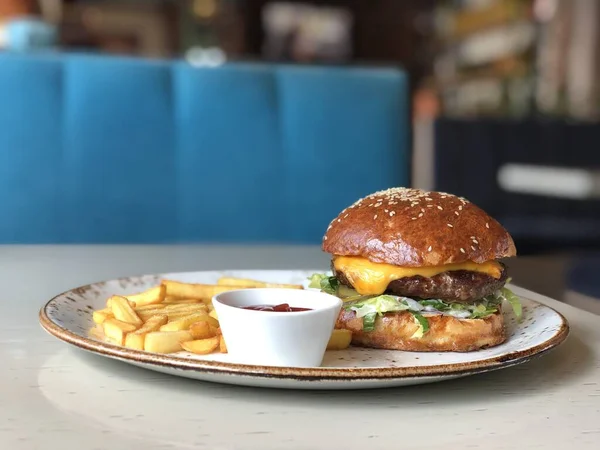 Taze iştah açıcı susamlı burger, beyaz bir tabakta, patates kızartmasıyla, küçük bir kavanozda domates ketçapıyla, kafeterya masasında. Yakın plan. Yan görünüm.