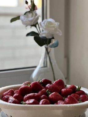 Taze olgun sulu çilekler beyaz bir pencere pervazında bir tabakta. Arka planda vazoda beyaz güller. 