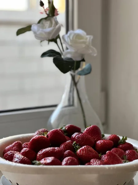Taze olgun sulu çilekler beyaz bir pencere pervazında bir tabakta. Arka planda vazoda beyaz güller. 