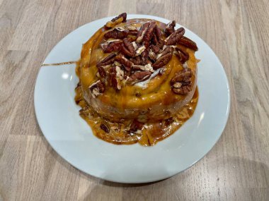 Üzerinde karamel sosu ve ahşap bir masada beyaz bir tabağa fındık serpiştirilmiş büyük, lezzetli bir tarçın rulosu. Yüksek kalite fotoğraf