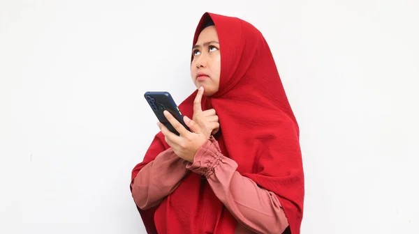 Asian woman in hijab confused and looking up to copy space while holding the smartphone