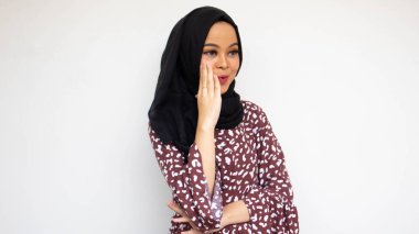 Kahverengi elbiseli, siyah tesettürlü, eli dudaklı, güzel bir Asyalı kadın. Gizli şeyler söylüyor, dedikodular fısıldıyor. Beyaz arka planda neşeli bir ifade var. İnsanlar İslam dini kavramları
