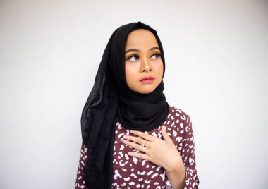 Mutsuz, tesettürlü genç Asyalı bir kadın beyaz arka planda yalnız, göğsü tutarken endişeli görünüyor.