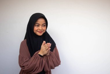 Türban takan Asyalı genç bir kadın yüzünde kocaman bir gülümsemeyle el sallıyor. Gri arka planda Endonezyalı kadın. Bayram / Bayram Fitr / Bayram El-Fitr selamlaması
