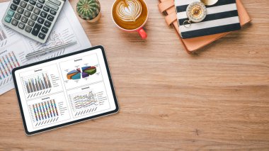 Tablet with charts and reports, calculator, pen, notebook and coffee on wooden desk workplace. Top view flat lay with copy space.	