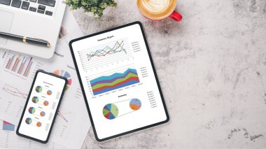 Tablet ve akıllı telefon, çizelgeler ve raporlar, hesap makinesi, kalem, defter ve kahve ofis masasında. Üst görünüm düzlüğü kopyalama alanı ile yatıyordu.