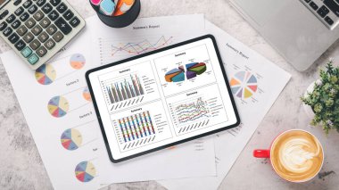Çizelge ve raporlarla dolu tablet, kalem, hesap makinesi, defter, fare ve klavye ofis masasında. Üst görünüm düzlüğü.