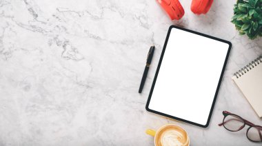 Boş ekran tableti, kulaklığı, kalemi, defteri ve kahvesi olan beyaz ofis masası çalışma alanı, fotokopi alanı olan üst görünüm dairesi..