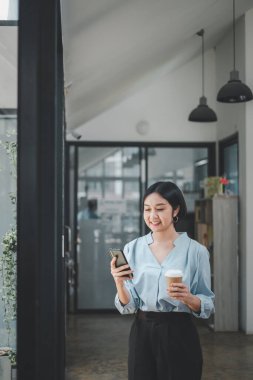 Attractive young woman talking on the smart phone and smiling while standing in office and looking smart phone.