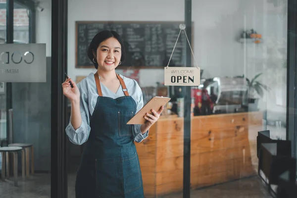Beautiful young barista woman in apron holding order paper and standing in front of the door of cafe with open sign board. Business owner startup concept.