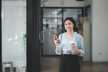 Attractive young woman talking on the smart phone and smiling while standing in office and looking smart phone.