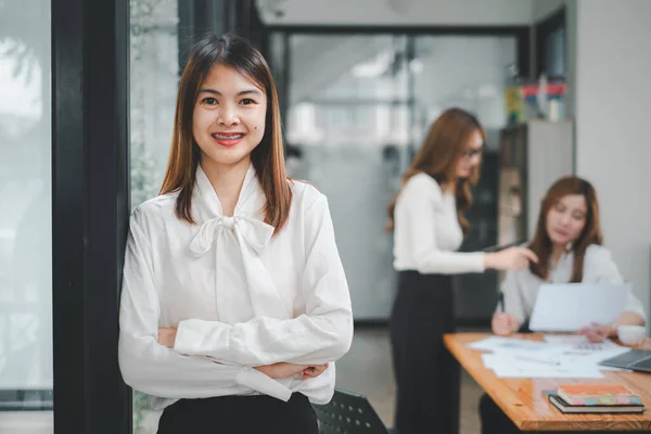 Happy business woman standing competently and smiling in open plan office with blurry collea gues sitting in office as background, Business concept.