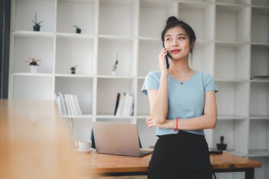 İş yerinde dizüstü bilgisayarına bakarken cep telefonuyla konuşan ve gülümseyen çekici genç bir kadın.