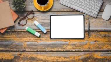 Boş ekran tableti, klavyesi, faresi, gözlüğü, kalemi, defteri ve kahvesi olan ahşap masa başı işyeri..