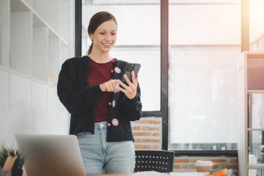 Attractive young woman talking on the smart phone and smiling while standing in office.