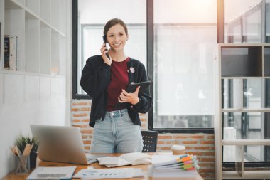 Attractive young woman talking on the smart phone and smiling while standing in office.