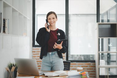 Attractive young woman talking on the smart phone and smiling while standing in office.