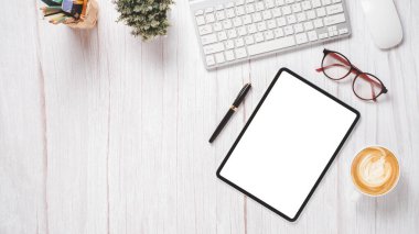 Boş ekran tableti, klavyesi, faresi, gözlüğü, kalemi ve kahvesi olan beyaz ahşap masa..