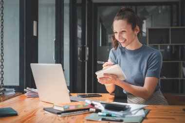 Ofis masasında oturan gülümseyen iş kadını müşterileri ya da iş arkadaşlarıyla bilgisayar başında konuşurken bir not defterine notlar yazıyor..