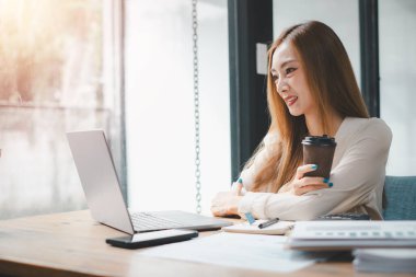 İş kadını kahve bardağını elinde tutuyor ve dizüstü bilgisayara bakıyor, işte bir gülümsemeyle mutluluk yaratıyor, yaratıcı ofiste çalışan Freelancer.