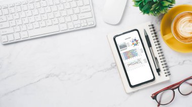 Ekran akıllı telefon ekranı ve ofis aletleri üzerinde özet grafik raporları olan beyaz ofis masası çalışma yeri.