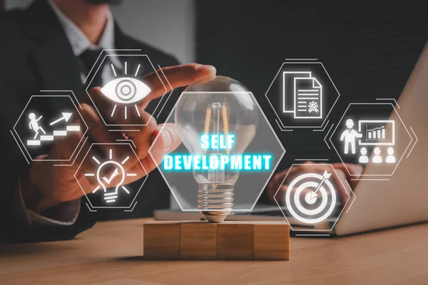 Self development concept. Businessman hand holding lightbulb on office desk with self development icon on virtual screen. Goals, Motivation, Training, Learning, Skill, Creativity, Critical Thinking.