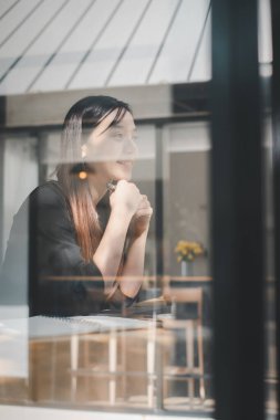 Düşünceli genç bir kadın pencereden dışarı bakıyor, masada bir defterin yansıması var.