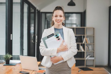 Gülümseyen profesyonel bir genç kadın, modern ofis ortamında evrakları tutuyor, kendine güveni tam..