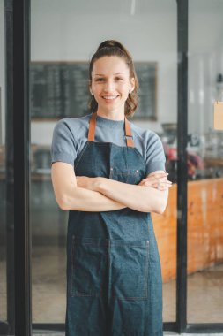 Kot önlüklü neşeli bayan kafe çalışanı kahve dükkanının önünde kollarını kavuşturmuş duruyor.