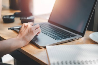 Bir insanın elinde kalem tutarken yakın plan görüntüsü, açık bir dizüstü bilgisayarın yanına yazmaya hazır, çoklu görev ve üretkenliği vurguluyor