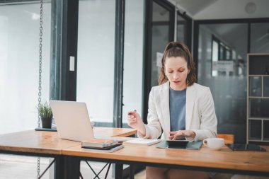 Odaklanmış iş kadını, iyi aydınlatılmış bir ofiste dizüstü bilgisayar ve kahvenin yanında belgeleri analiz ediyor.