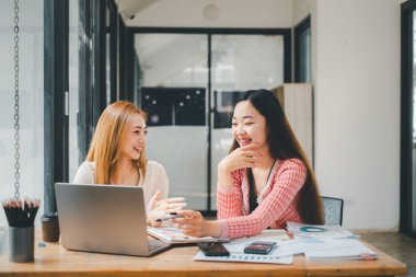 İş ekibi analizi konsepti, iki gülümseyen bayan meslektaşın bir dizüstü bilgisayar ve masadaki belgelerle dostane bir sohbete girişmesi.