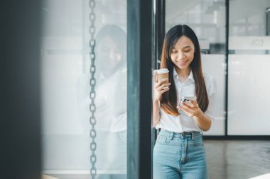 Ofis ortamında elinde kahve fincanı tutarken akıllı telefonunu kullanarak günlük iş kıyafetleri içinde gülümseyen genç bir kadın.