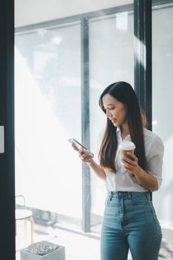 İş gevşeme konsepti güneşli bir şirket ortamında kahve molası verirken akıllı telefonunu kullanan gülümseyen profesyonel bir kadın.