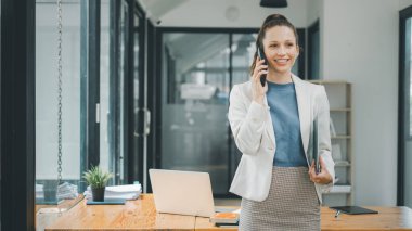 İş kadını konsepti, gülümseyen genç iş kadını dizüstü bilgisayarını iyi aydınlatılmış modern bir ofiste tutarken telefonla konuşuyor.