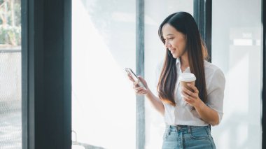 İş teknolojisi konsepti, rahat profesyonel kadın akıllı telefonunu kontrol ediyor ve modern ofis ortamında bir fincan kahvenin tadını çıkarıyor.