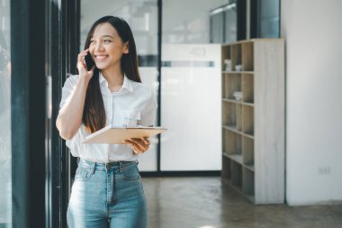 İş teknolojisi konsepti, neşeli genç bir iş kadını elinde parlak bir ofiste bir pano tutarken bir telefon görüşmesi yapıyor.